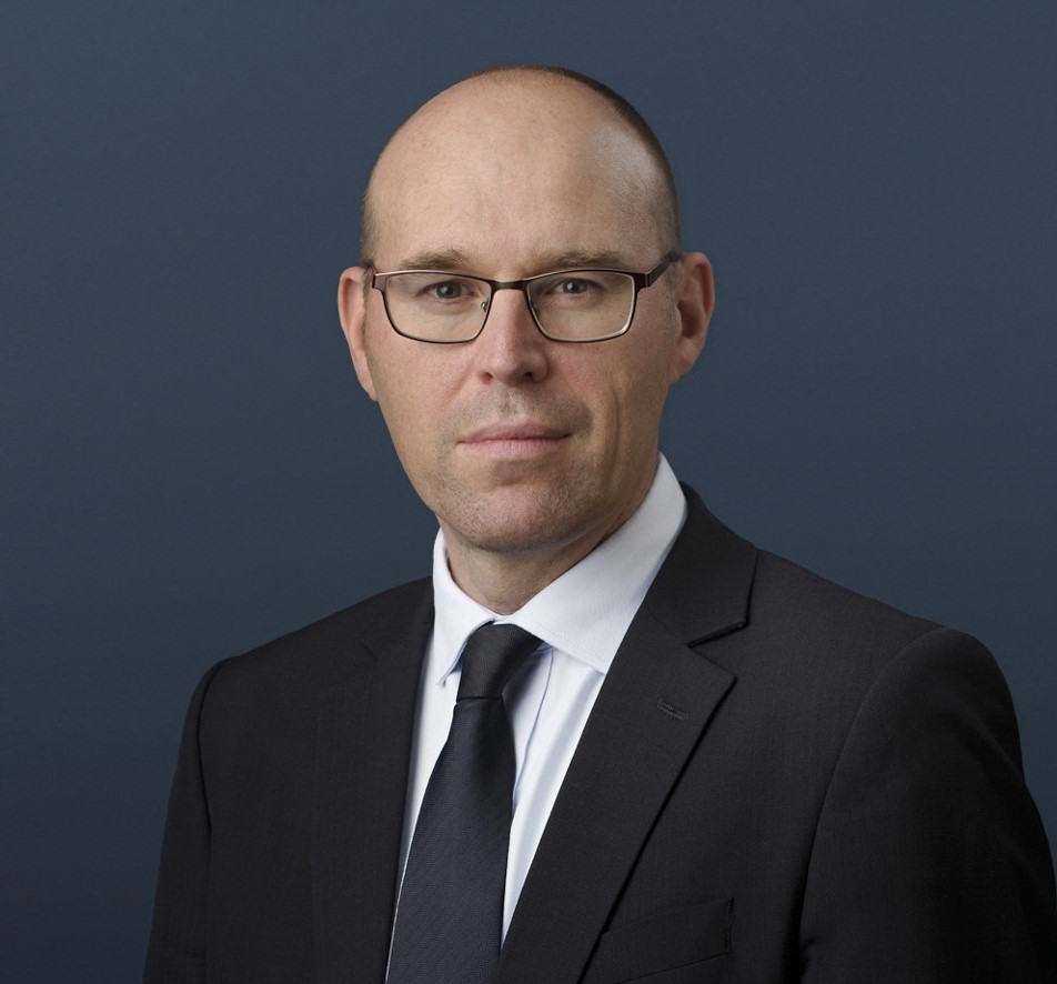 The image shows a head and shoulders photo of Simon Lewis. The person is against a blue background and is wearing a navy blue suit, looking to camera